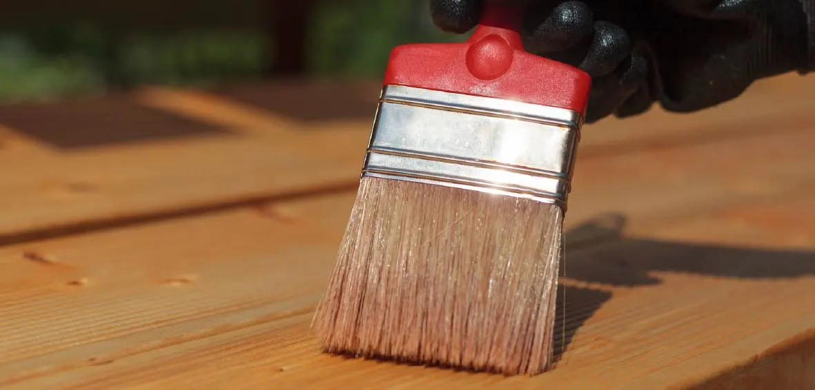 A close-up of a wide paint brush applying paint stripper to wood decking.