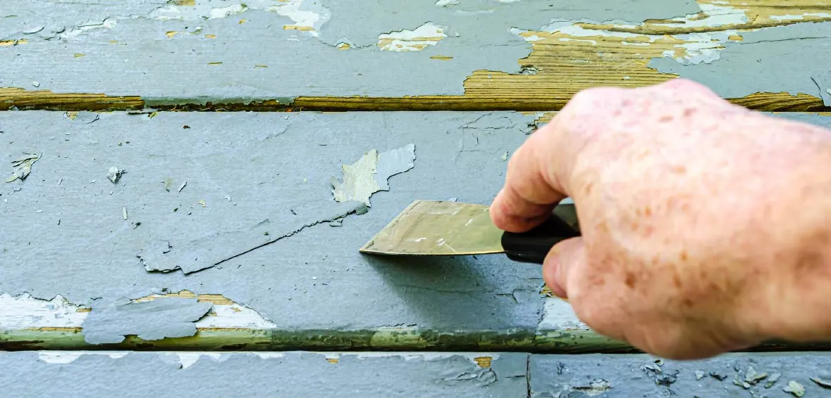 A close-up of someone using a paint scraper to manually removing blue paint from an old wood deck.