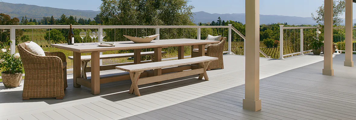 Photo of a partially covered light gray TimberTech deck with white railing and vertical cable infill. A long wooden table with matching benches and wicker arm chairs has a bottle of wine and glasses set atop it, as if ready to entertain a small crowd.