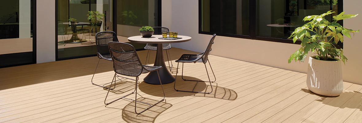 Modern home with floor to ceiling glass doors and large windows has a tan colored deck with a circular table and four chairs around it a few feet from a large potted plant.