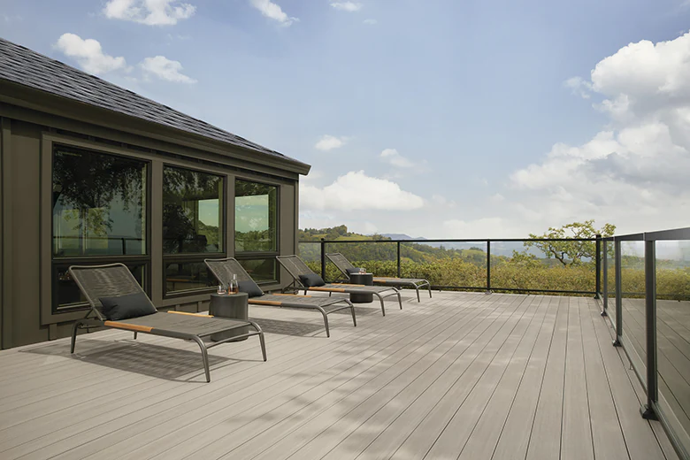 Four lounge chairs sit in a row on a modern light tan deck with a glass infill railing around the perimeter