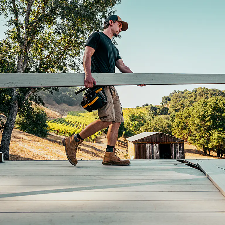 A TimberTech contractor walks with a light tan deck board at a job site next to a vineyard