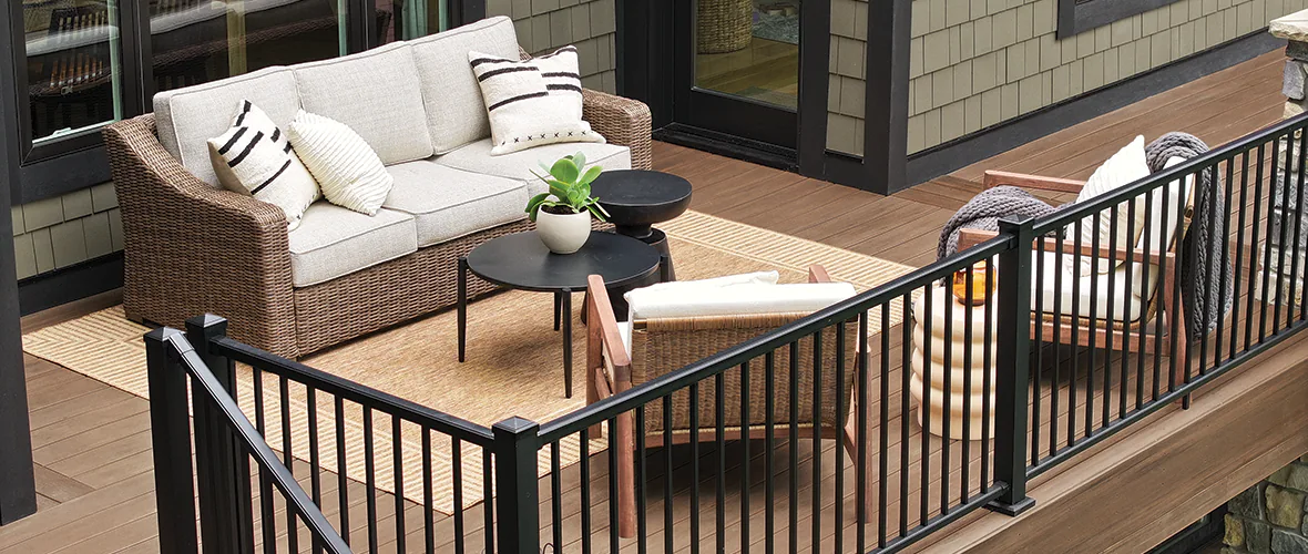 A second story deck built with richly-hued medium brown deck boards and a black railing. Wicker furniture with taupe colored pillows area present creating a seating area that emphasizes the use of natural fibers.
