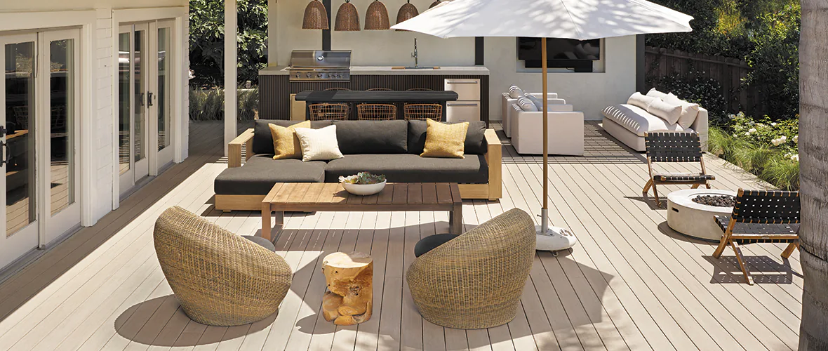 A light tan deck extends from a white house. French doors lead to the outdoor living room and kitchen area of the deck. Three seating areas are visible with an array of chairs and styles.