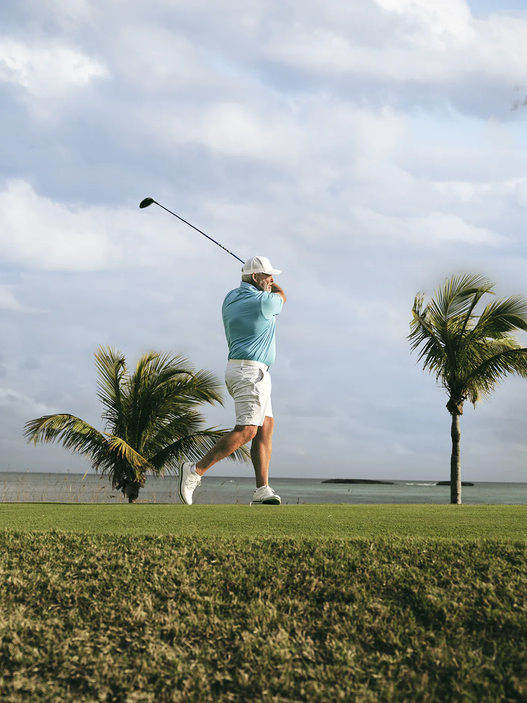 Darren swings his golf club on the grass at a local golf course. Two tropical trees in the background frame the photo on either side.