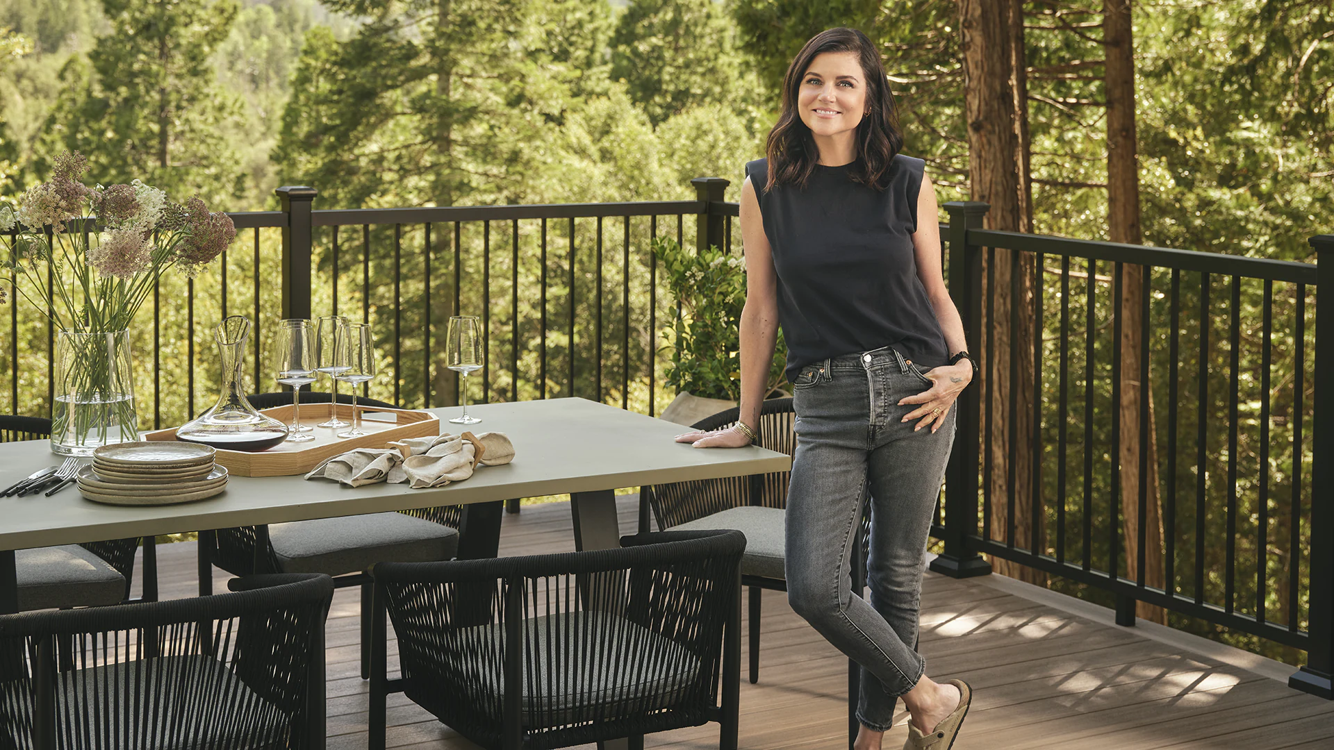 Tiffani Thiessen stands next to an outdoor dining room table on her deck, her legs crossed while smiling and gently leaning towards the dining set up.